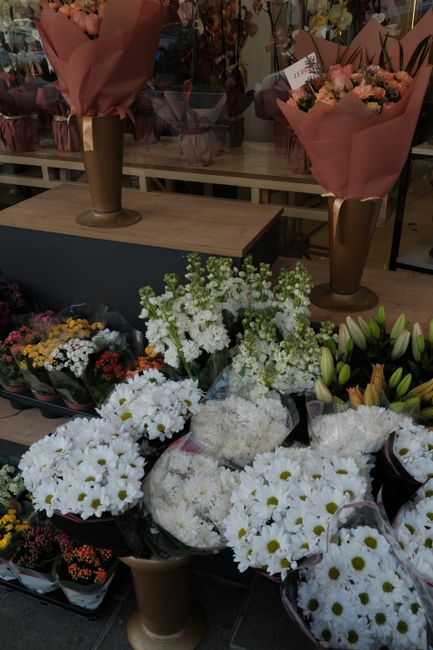 An arrangement of fresh white daisies and green foliage in black wrapping, placed on a table in a flower shop. Behind the daisies, there are other floral displays including bouquets of pink and peach 