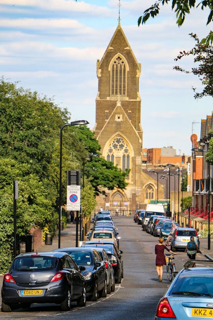 The image depicts a narrow street in East Finchley lined with parked cars on both sides, leading towards a historic church with a tall, pointed steeple and large arched stained glass windows. The stre
