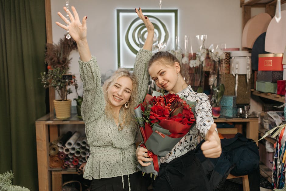 Two young women in a floral workshop or flower shop, one holding a large bouquet of deep red roses wrapped in dark grey and red paper, while the other cheerfully raises her hands in celebration. The b